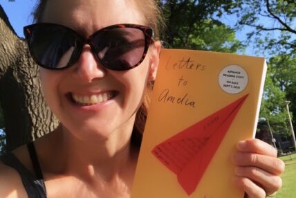 Author Lindsay Zier-Vogel, white woman with large dark sunglasses holding up a yellow book with a red paper airplane with the title Letters to Amelia